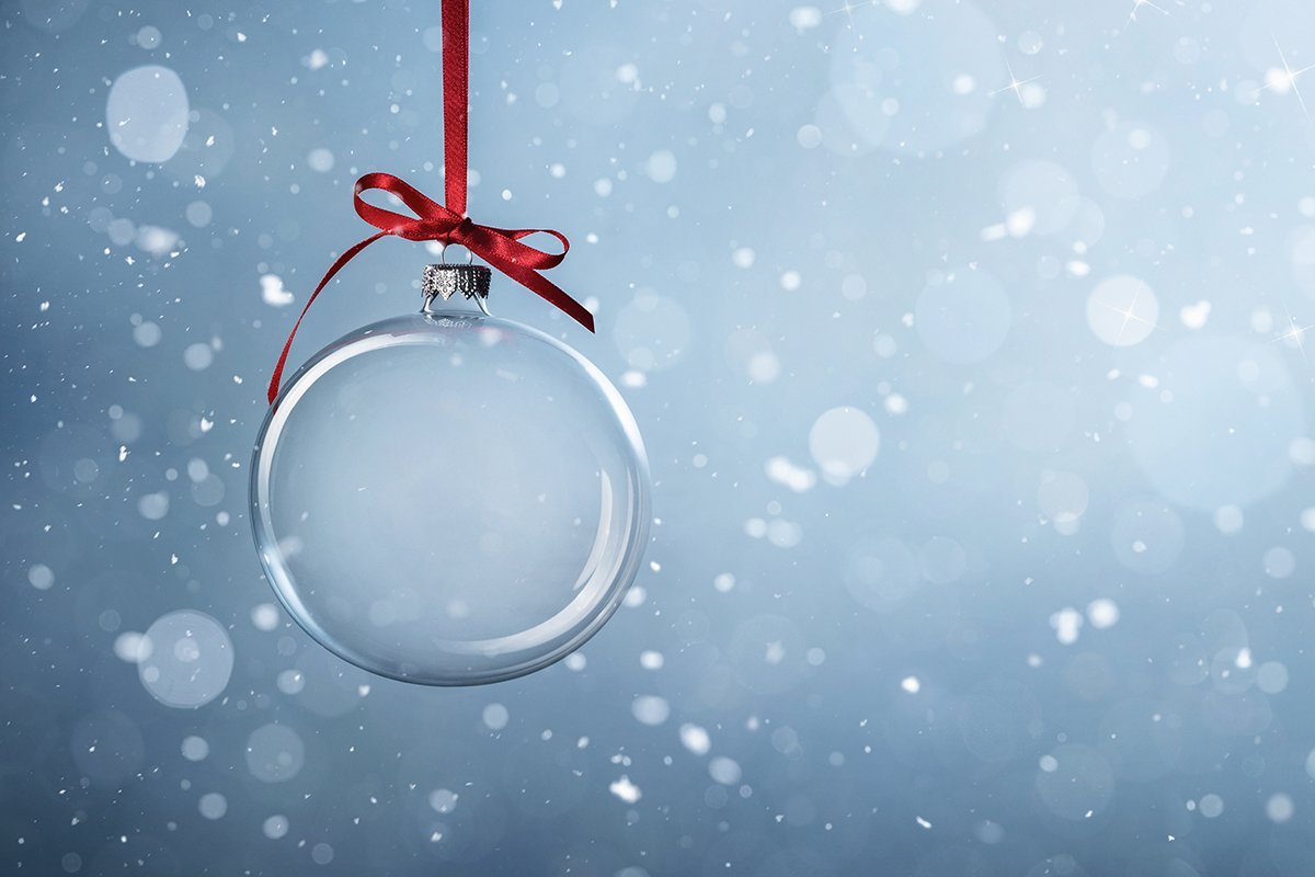 Eine durchsichtige Glas-Weihnachtskugel hängt an einer roten Stoffschleife; der Hintergrund ist ein sanftes Hellblau mit kleinen weißen Punkten und Flocken..