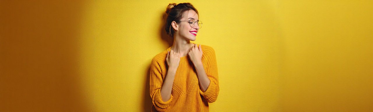 Junge Frau mit Brille und hochgesteckten Haaren trägt einen gelben Strickpullover und lächelt verträumt zur Seite. Sie steht vor einer gelben Wand.