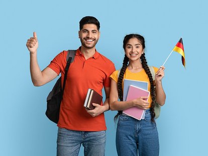 Ein junger Mann und eine junge Frau stehen nebeneinander vor einem hellblauen Hintergrund. Beide lächeln, tragen Rucksäcke und halten Bücher. Die Frau hält zusätzlich eine kleine deutsche Flagge.