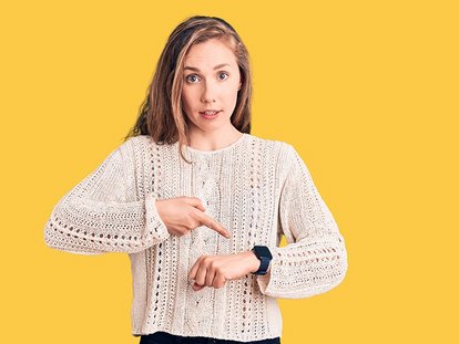 Junge Frau mit langen Haaren und hellem Strickpullover zeigt mit dem Finger auf ihre Smartwatch am Handgelenk. Sie schaut überrascht oder fragend in die Kamera. Hintergrund ist einfarbig gelb.