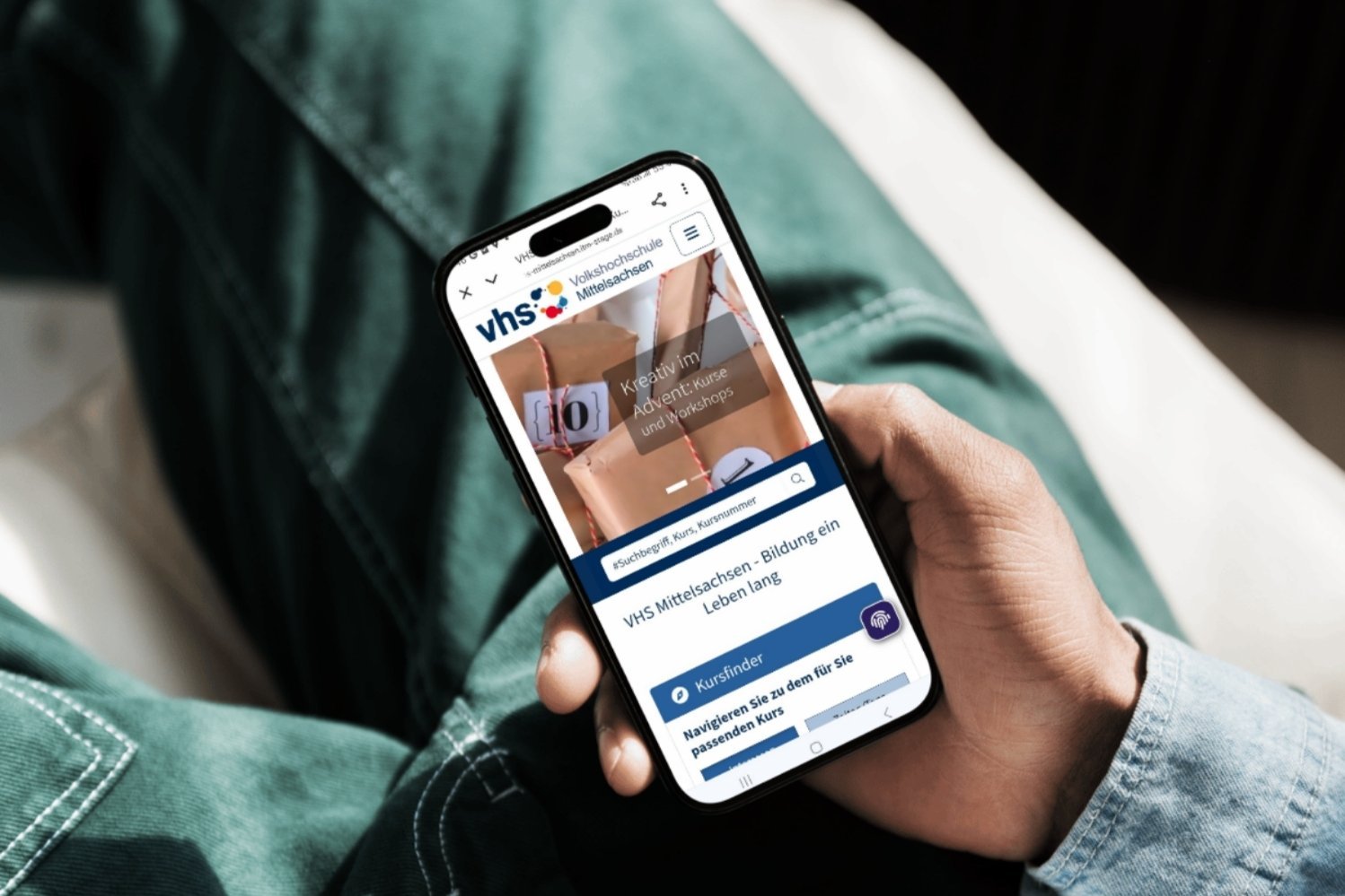Eine Person hält ein Smartphone in der Hand, auf dem die Website einer Volkshochschule geöffnet ist. Auf dem Bildschirm ist die Startseite mit einem Kursfinder und dem Text „Kreativ im Advent – Kurse und Workshops“ zu sehen.