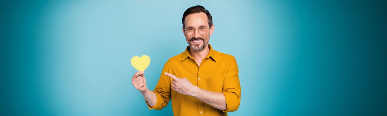Mann mittleren Alters mit Brille und Bart hält ein gelbes Papierherz in der Hand und zeigt mit dem Finger darauf. Er trägt ein senfgelbes Hemd und lächelt. Hintergrund ist einfarbig hellblau.